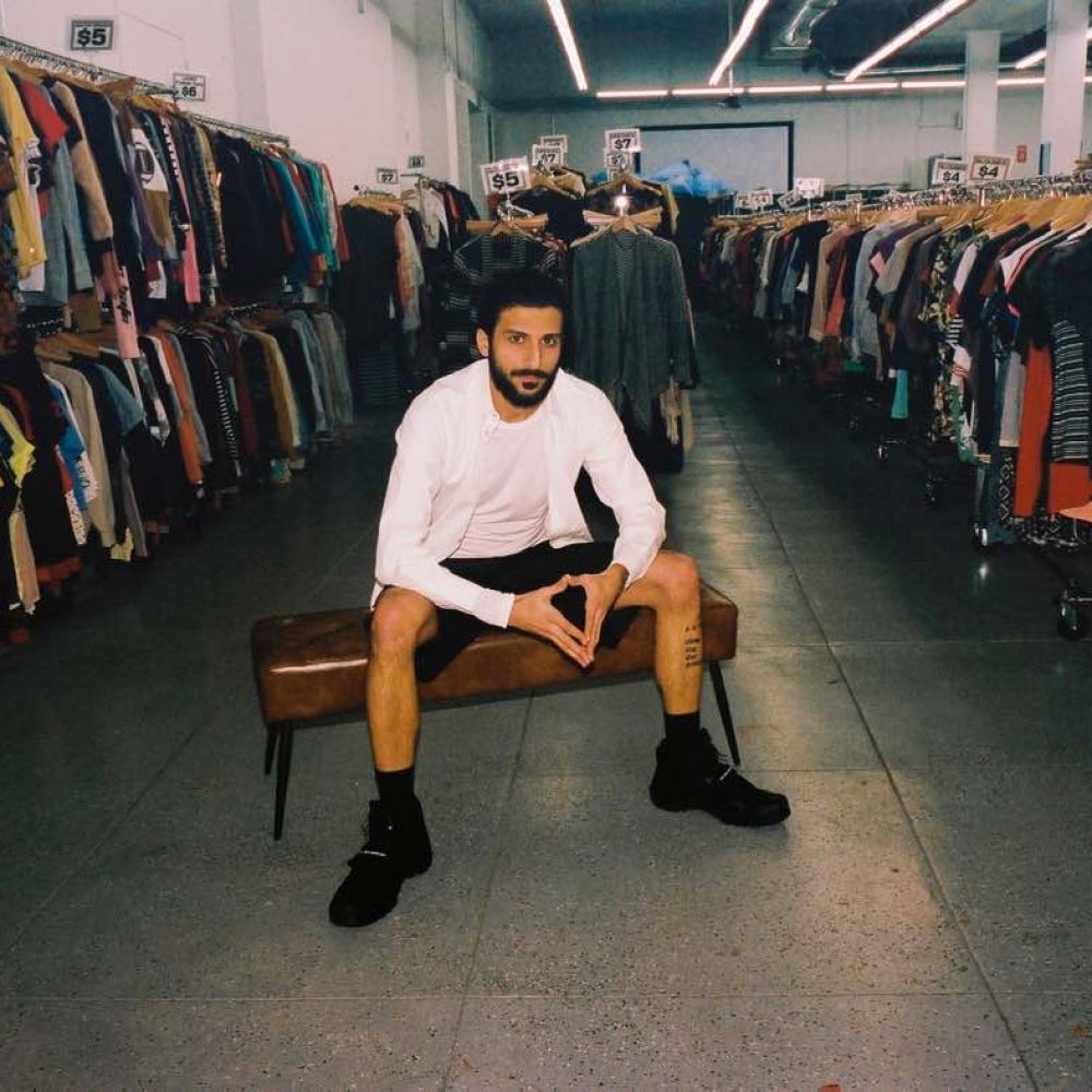 Christopher Ohanian poses sitting down in between racks in his new thrift store Public Estrellas.